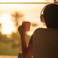 Rear view of woman with headphones and cup of coffee sitting on chair near window and looking on palm trees